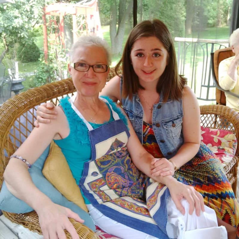 Two women, one older with glasses and a blue top, and a younger woman with a denim vest, sit on a wicker couch with a colorful patterned blanket, while a lush green, tree-filled background is visible through an open porch.