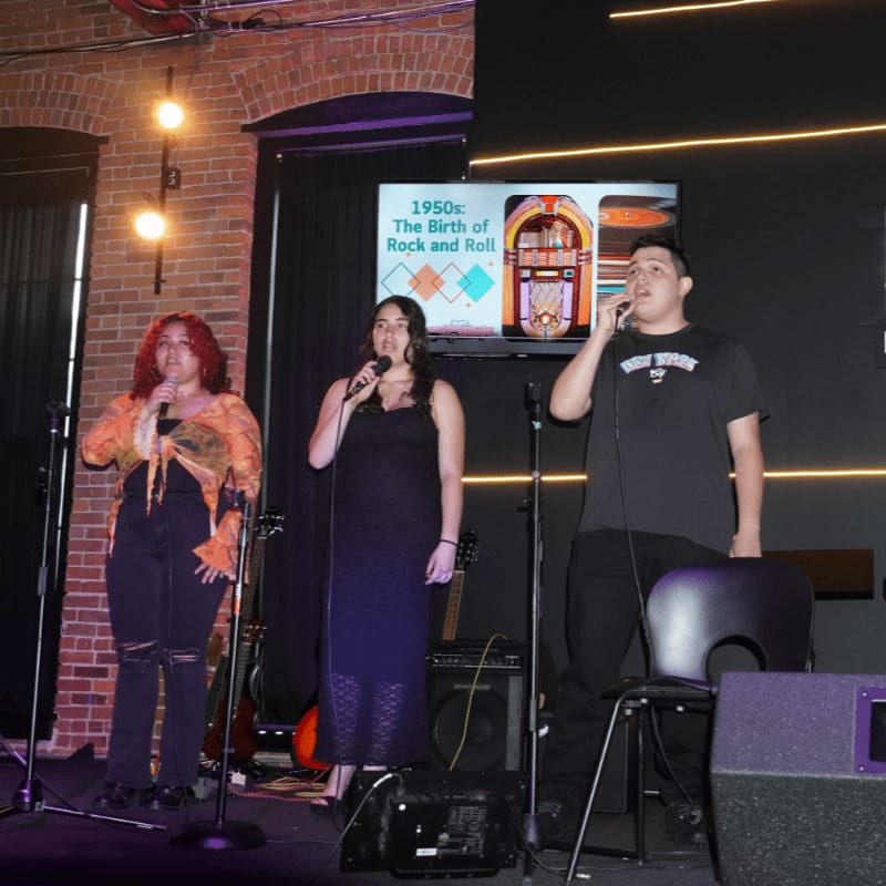 Three young people sing into microphones on a stage with musical instruments in the foreground, while a screen displaying "1950s: The Birth of Rock and Roll" with a jukebox is visible in the background.