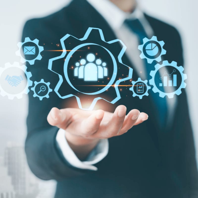 A businessman in a suit holds out his hand with glowing gears representing teamwork and business processes against a blurred city background.