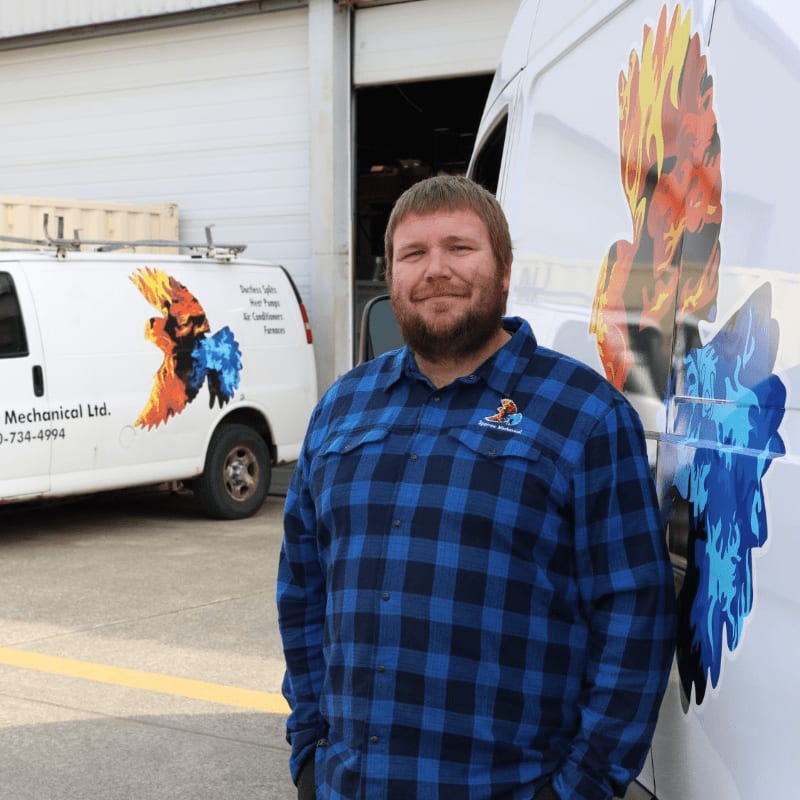 A man in a blue plaid shirt stands in front of a white van with a fiery bird logo, while another similar van is parked behind him.