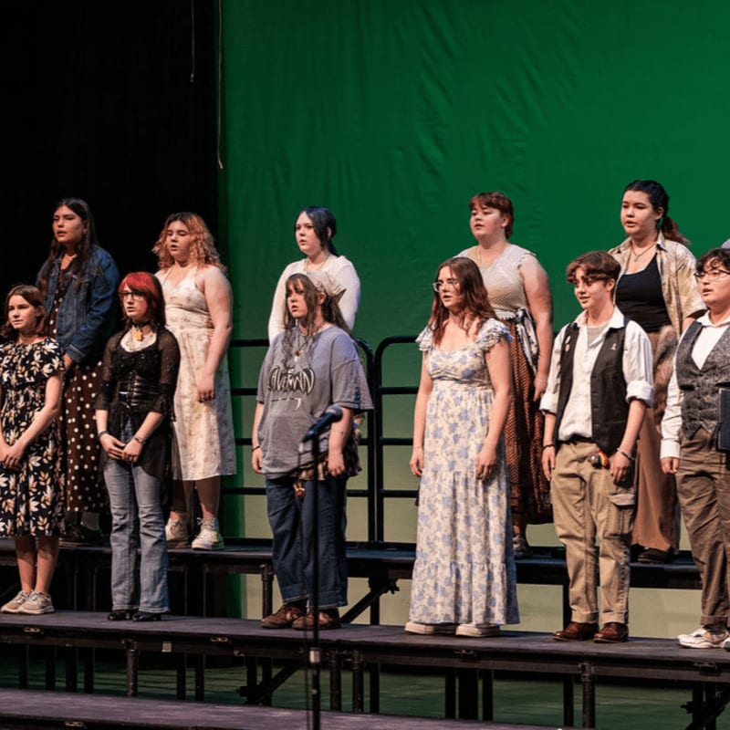 A group of young people stand on risers in the foreground, singing before a green backdrop in the background.