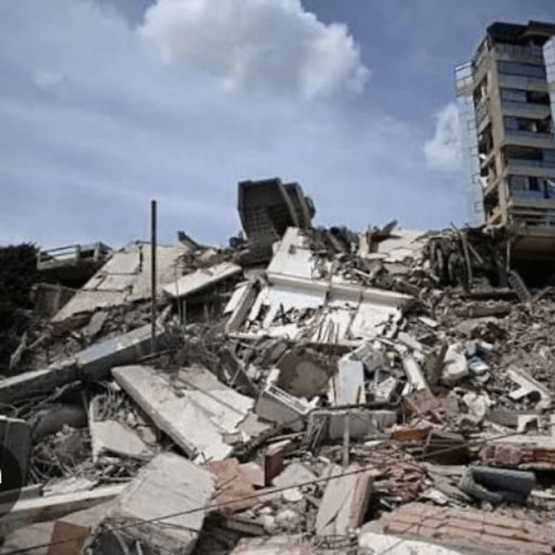 A massive pile of rubble from a collapsed building dominates the foreground, with a partially standing, damaged skyscraper visible in the background under a cloudy sky.