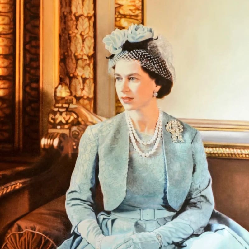 A young Queen Elizabeth II, adorned in pearls and a light blue suit with a matching hat, sits regally on a brown couch in front of a richly decorated golden wall.