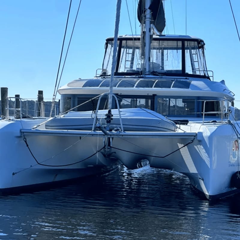 A white catamaran with a dinghy on its deck rests on calm water in front of a clear blue sky.