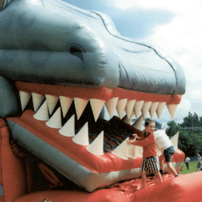 Two children play on a large, inflatable dinosaur with a bright red mouth and sharp white teeth, set against a backdrop of a cloudy sky and distant trees.