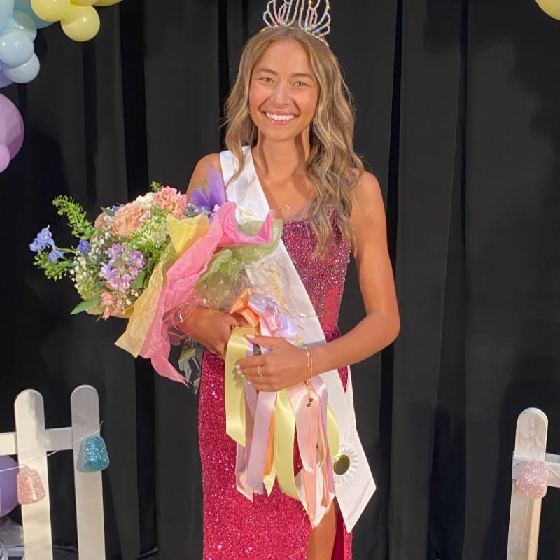 A smiling young woman in a sparkly pink dress and a tiara holds a bouquet of flowers in front of a black curtain adorned with balloons and a white picket fence.