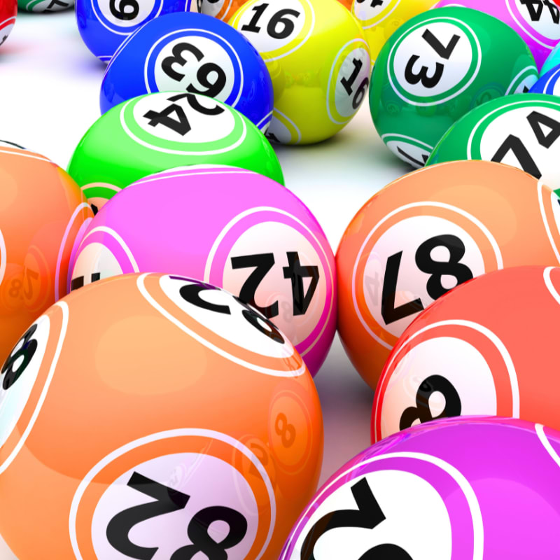 Colorful lottery balls with numbers are scattered across a white surface, with more balls visible in the blurred background.