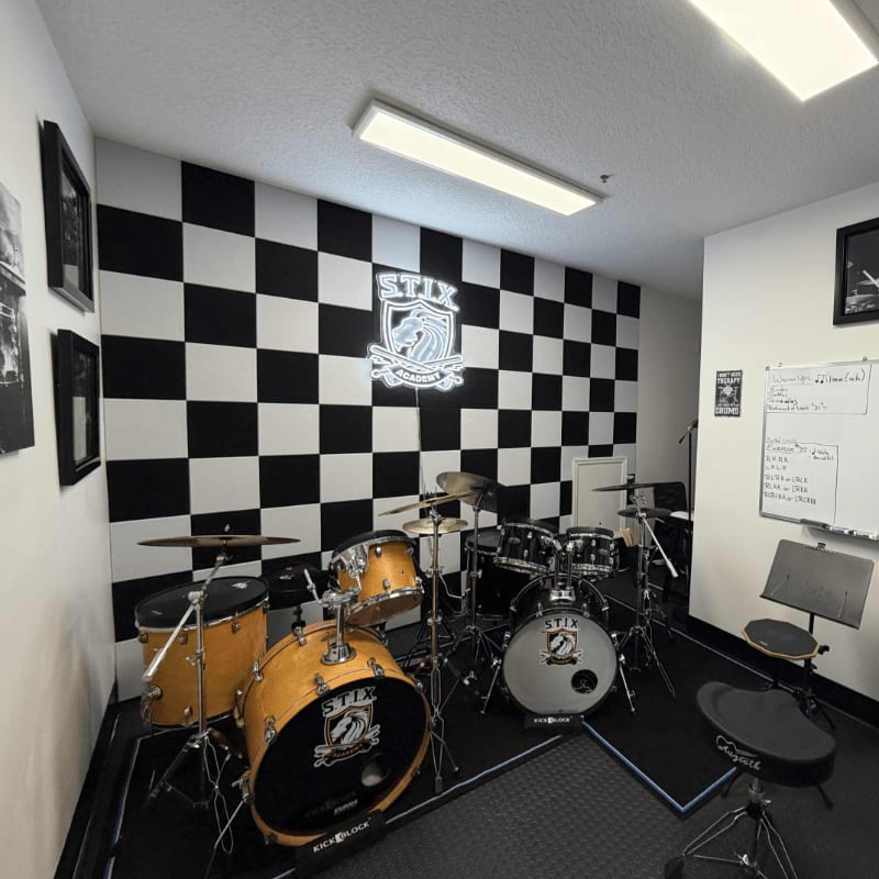A drum set with a checkered wall and a logo in the background is in the foreground.