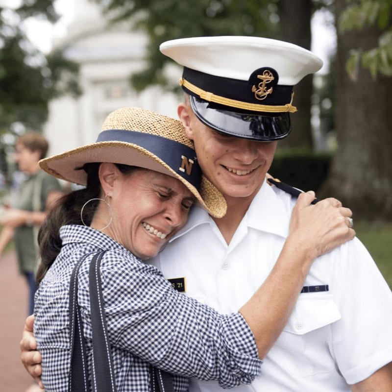 A woman in a straw hat hugs a man in a naval uniform, with a blurred building and trees in the background.