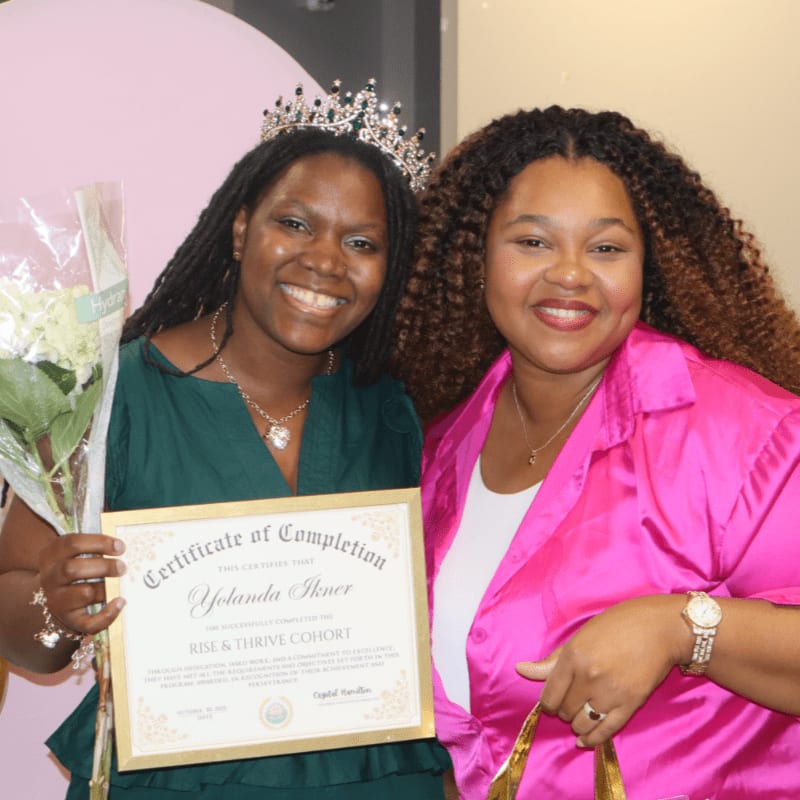 Two smiling women pose for a photo, one wearing a tiara and holding a certificate and flowers, with a pink circular backdrop behind them.