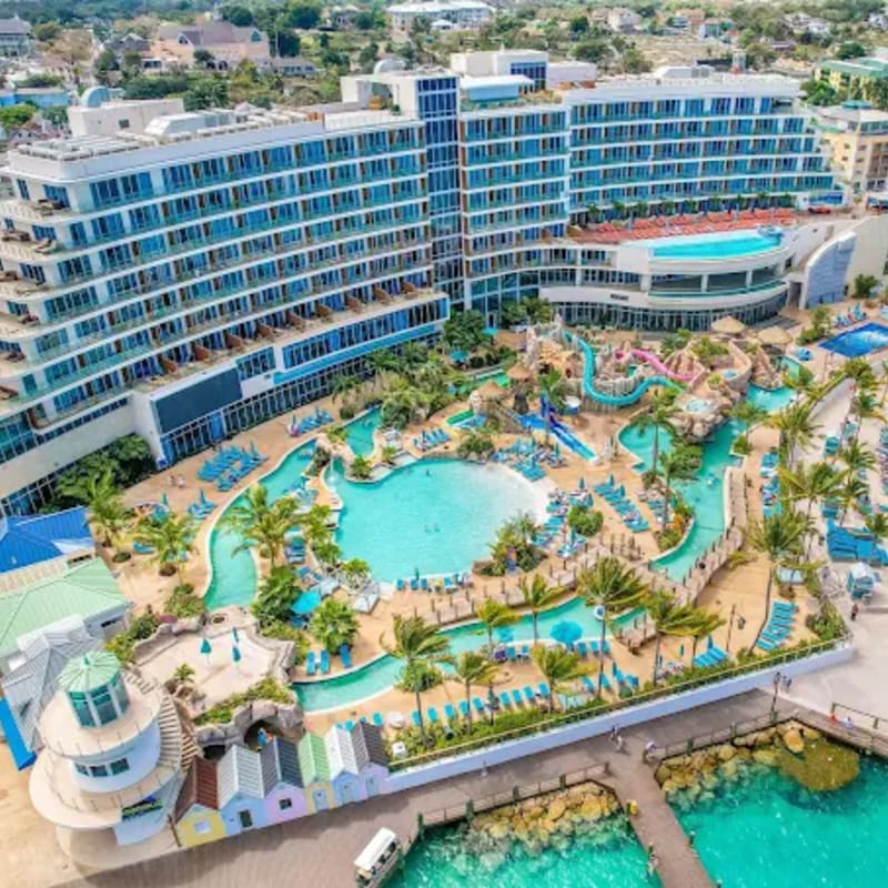 A sprawling resort with a large water park and pool complex in the foreground is set against a backdrop of a coastal town with buildings and palm trees.