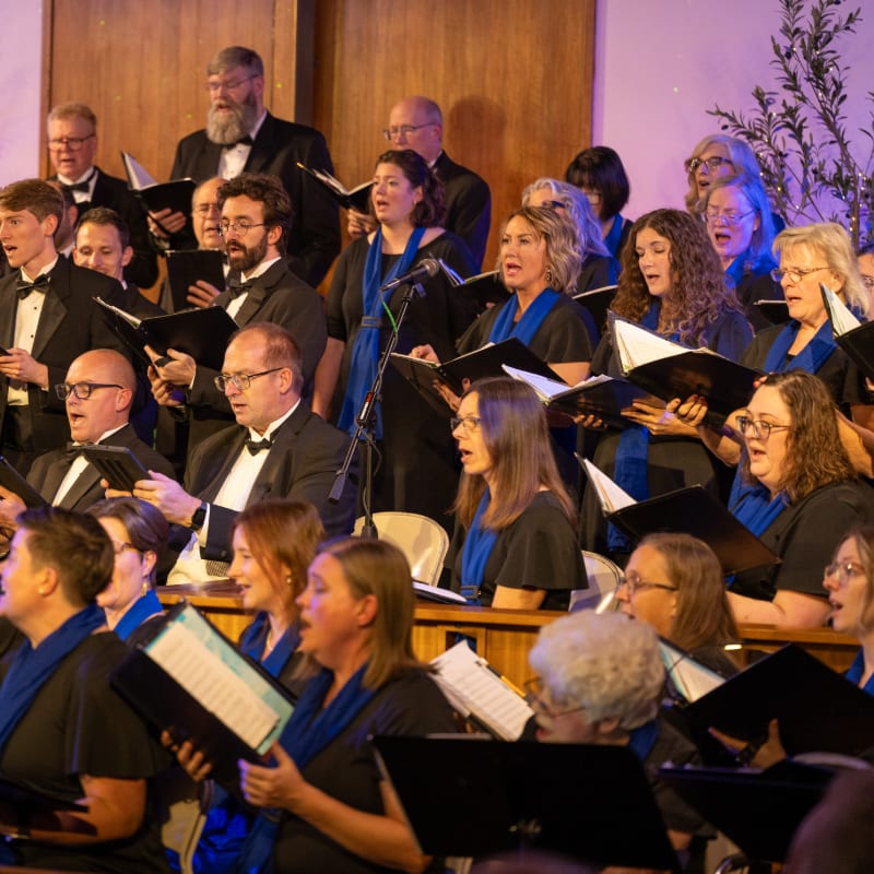 A choir of men and women in black attire with blue scarves sing from sheet music in front of a wooden wall.