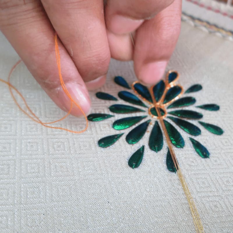 Fingers guide an orange thread through a needle, stitching a metallic green floral design onto a textured white fabric.