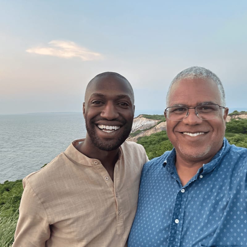 Two smiling men pose for a selfie with a scenic ocean and cliffside view in the background.