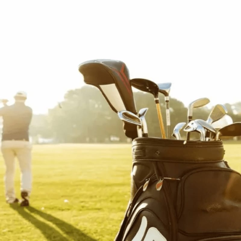 A golf bag filled with clubs stands in the foreground on a sunny golf course, with a golfer swinging in the blurred background.