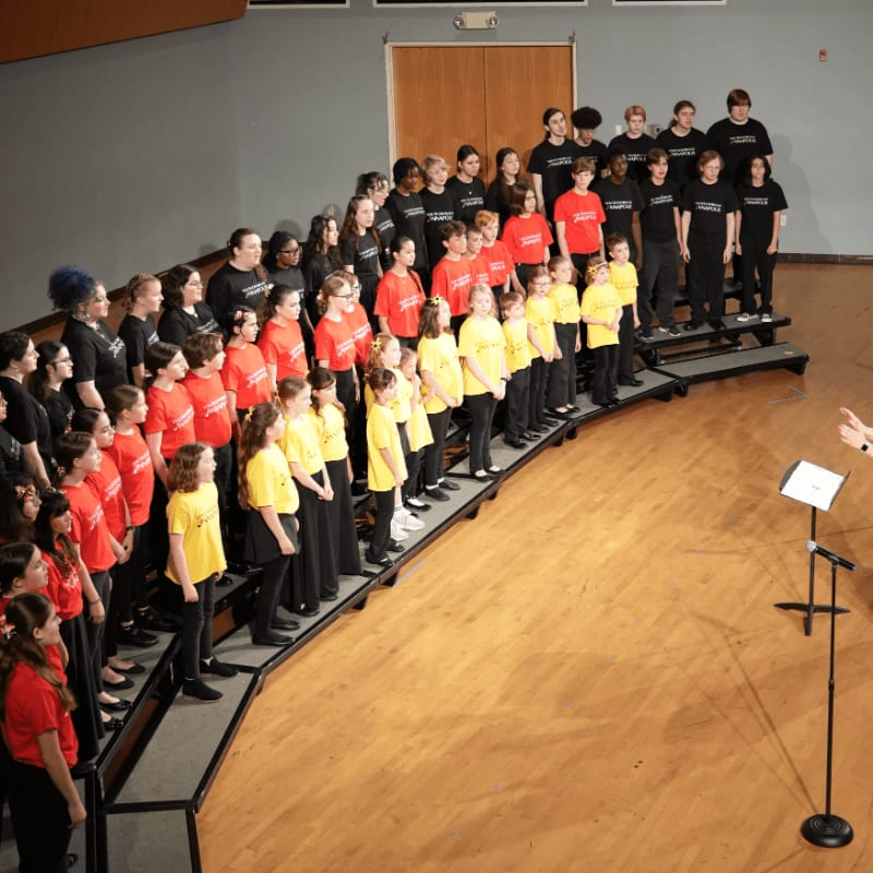 A choir of young people in colorful shirts stands on risers in a room with a wooden floor and a plain wall in the background.