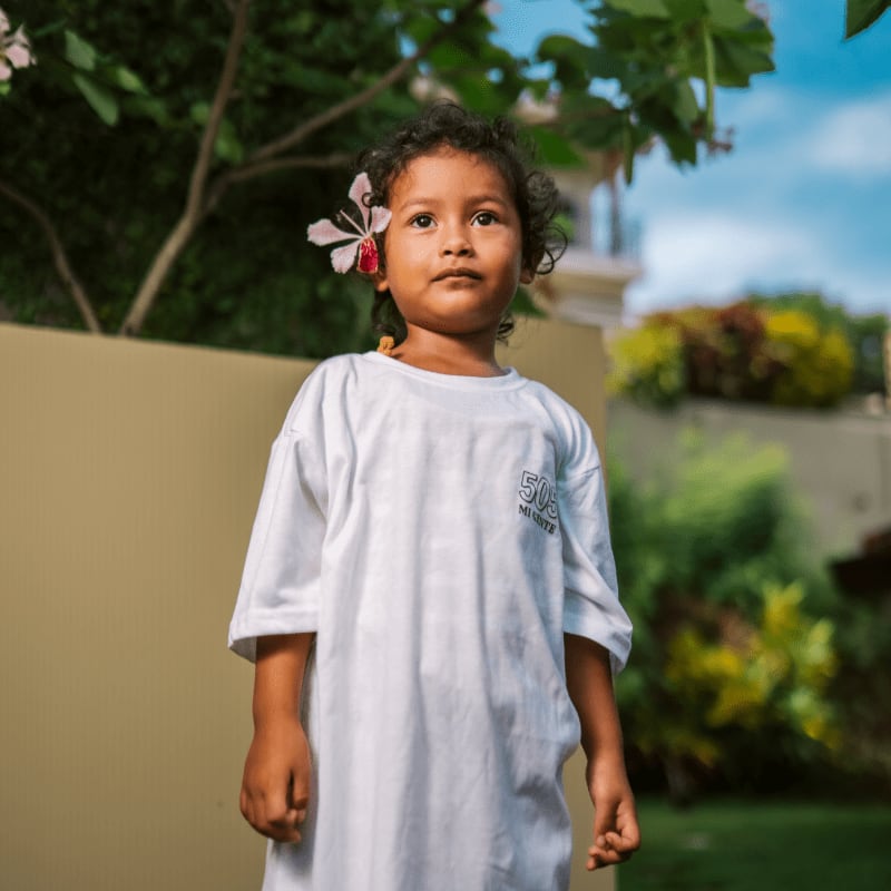 A young child with a flower in their hair stands in front of a beige wall, with lush greenery and a bright blue sky in the background.