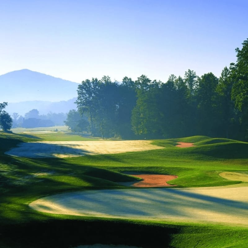 Rolling green fairways with sand traps in the foreground lead to a tree-lined course with a hazy mountain in the background under a clear blue sky.