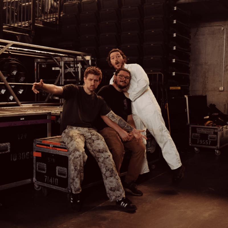 Three men pose in front of stacked equipment cases and stage lighting.