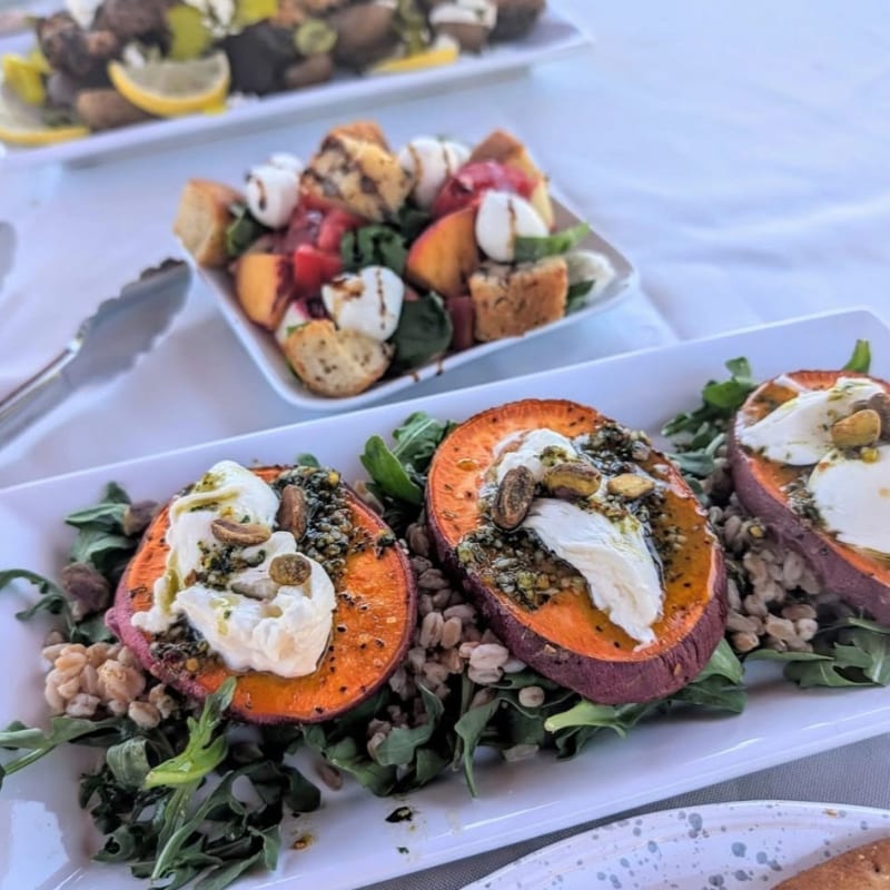 Roasted sweet potato halves topped with cheese and pistachios sit on a bed of greens and grains in the foreground, with a caprese salad in the blurred background.