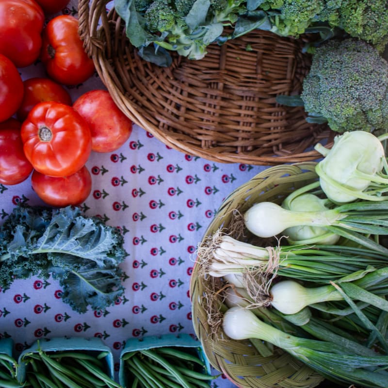 Fresh tomatoes and kale lie on a patterned cloth in the foreground, while baskets of broccoli and green onions fill the background.