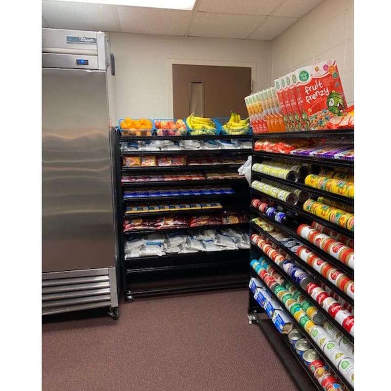 A stainless steel refrigerator stands to the left of shelves stocked with packaged snacks and canned goods, with a doorway visible in the background.