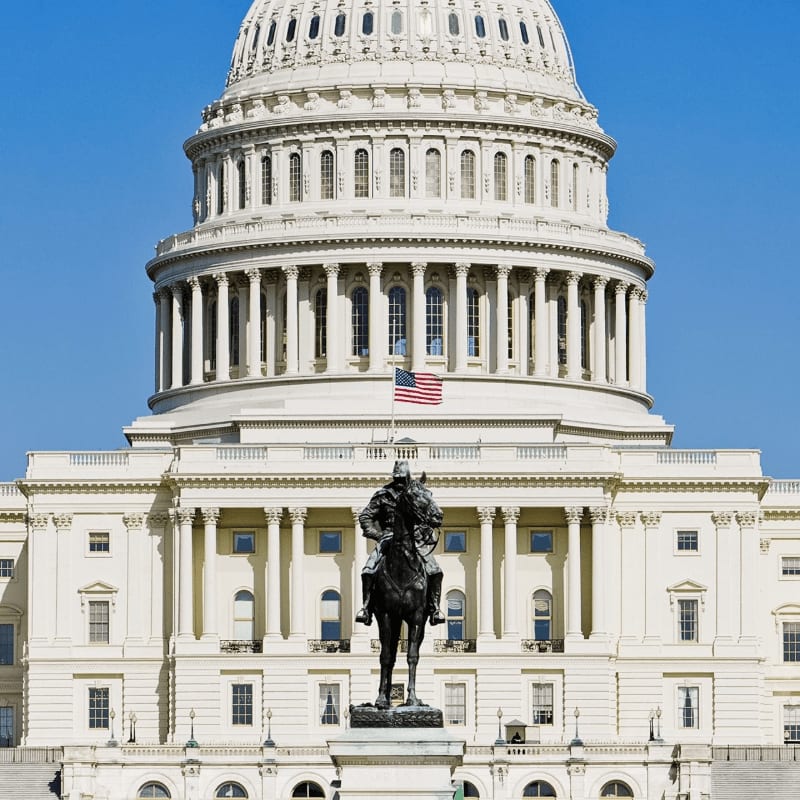 A bronze equestrian statue stands before the grand, white dome of the U.S. Capitol building under a clear blue sky.
