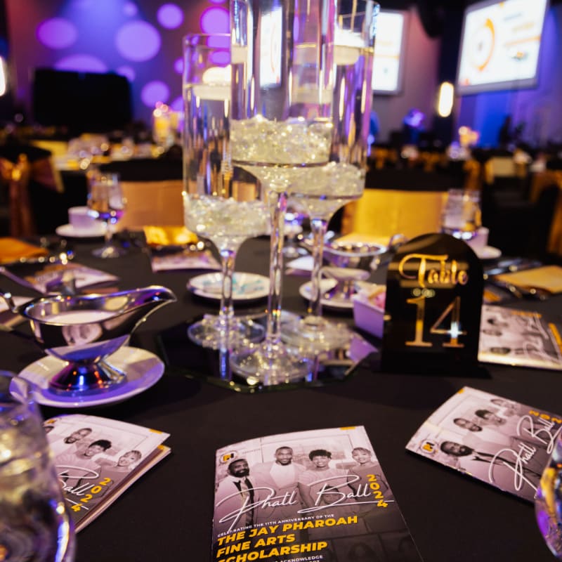 A formal table setting with a centerpiece of tall vases filled with water and crystals is in the foreground, while a dimly lit banquet hall with purple and gold decor is in the background.
