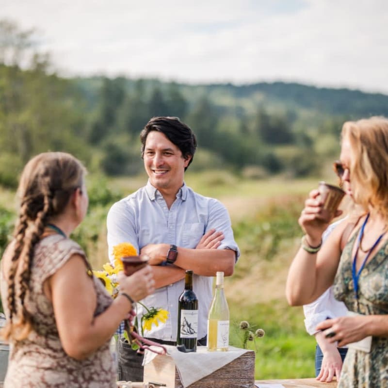 Three people converse at a table with wine bottles in the foreground, while a blurred, green, hilly landscape stretches into the background under a bright sky.