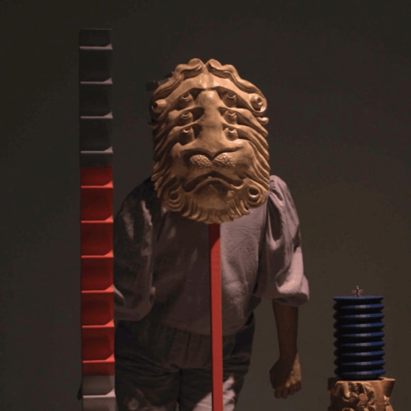 A wooden lion mask adorns a figure in a grey shirt, standing before a dark background with abstract sculptures of stacked blocks and a blue cylindrical object.