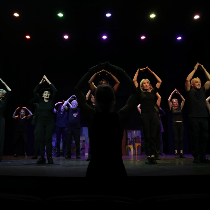 A group of performers in black outfits form a circle with their arms on a dimly lit stage with colorful lights overhead.