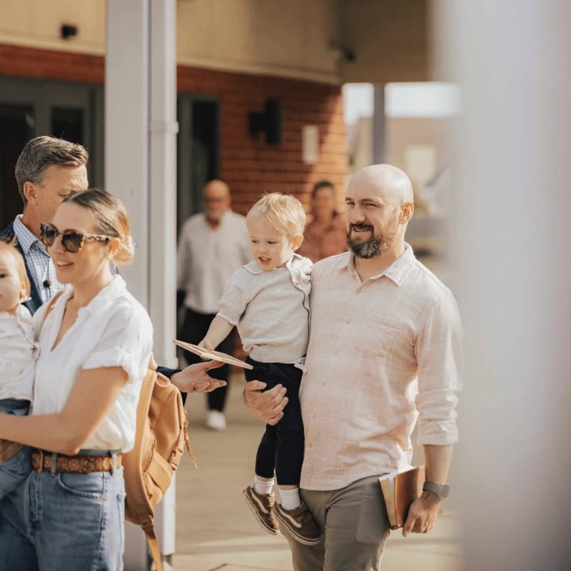 A smiling man holds a toddler while walking past a woman with sunglasses and a backpack, with other people and a brick building in the background.