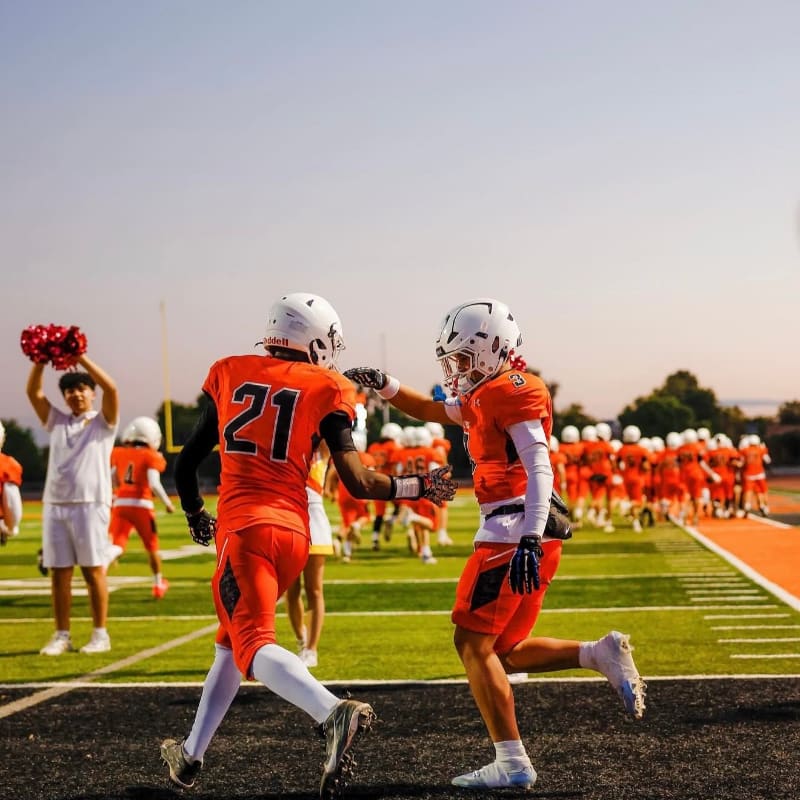 Two football players in orange jerseys high-five on a field with a cheering crowd in the background.