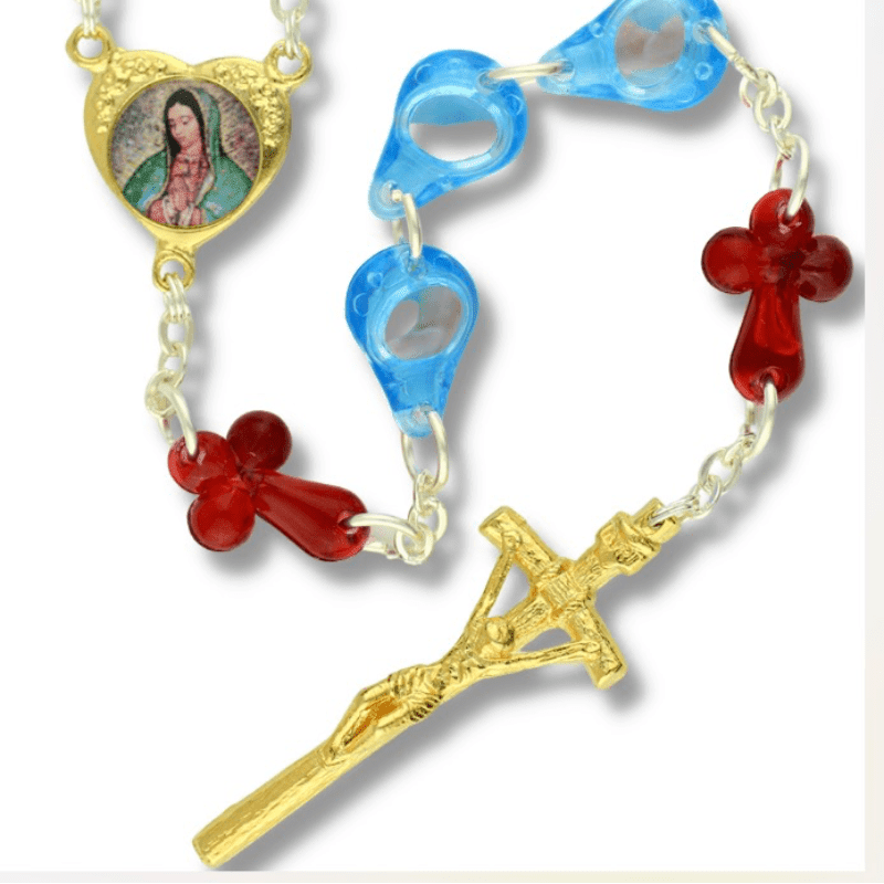 A gold crucifix and red cross beads are strung on a chain with blue oval beads and a heart-shaped medal of Mary in the foreground, against a plain white background.