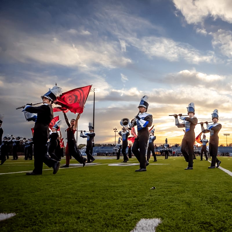 A marching band performs on a green field with a dramatic sunset sky in the background.