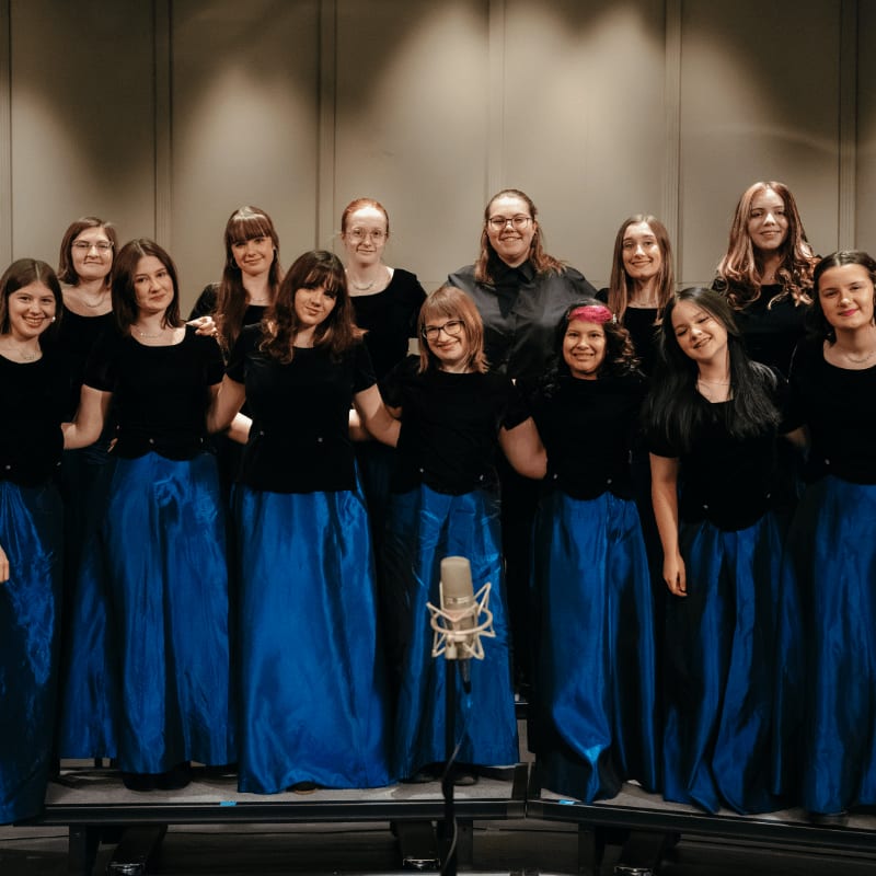 A group of young women in blue skirts and black tops stand in front of a textured wall, with a microphone in the foreground.