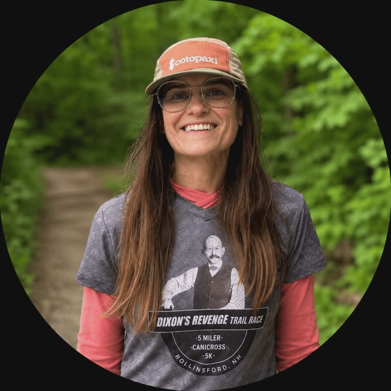 A smiling woman wearing a hat and a t-shirt with a trail race logo stands on a path in a wooded area.