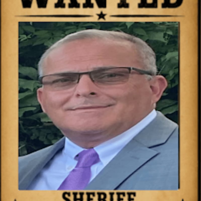 A man in a suit and tie is pictured on a wanted poster with a leafy green background.