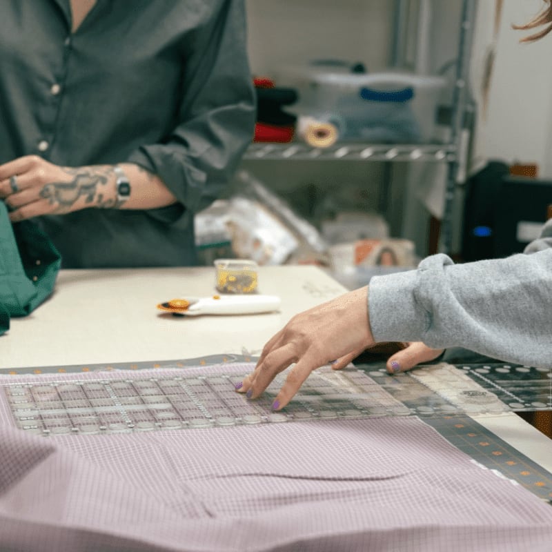 A person's hands are using a ruler to measure fabric in the foreground, while another person stands behind them in the background with shelves of supplies.