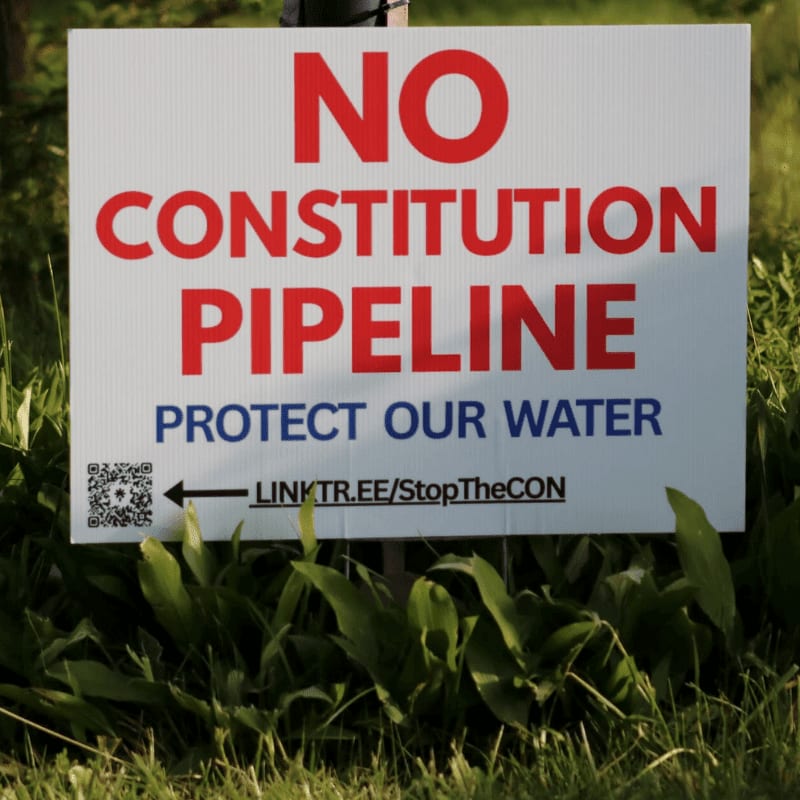 A white sign with red and blue lettering reading "NO CONSTITUTION PIPELINE PROTECT OUR WATER" is planted in green grass, with a blurred background of more grass and a fence post.