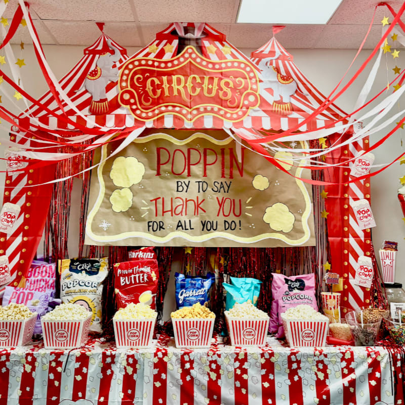 A circus-themed display features bags and buckets of popcorn in the foreground, with a large "CIRCUS POPPIN THANK YOU" banner and red and white striped tents in the background.