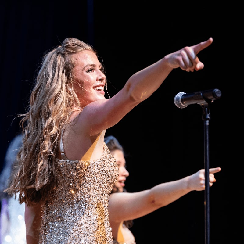A woman in a sparkling gold dress points forward on a stage with a microphone in front of her, while another woman in a similar dress stands behind her against a dark background.