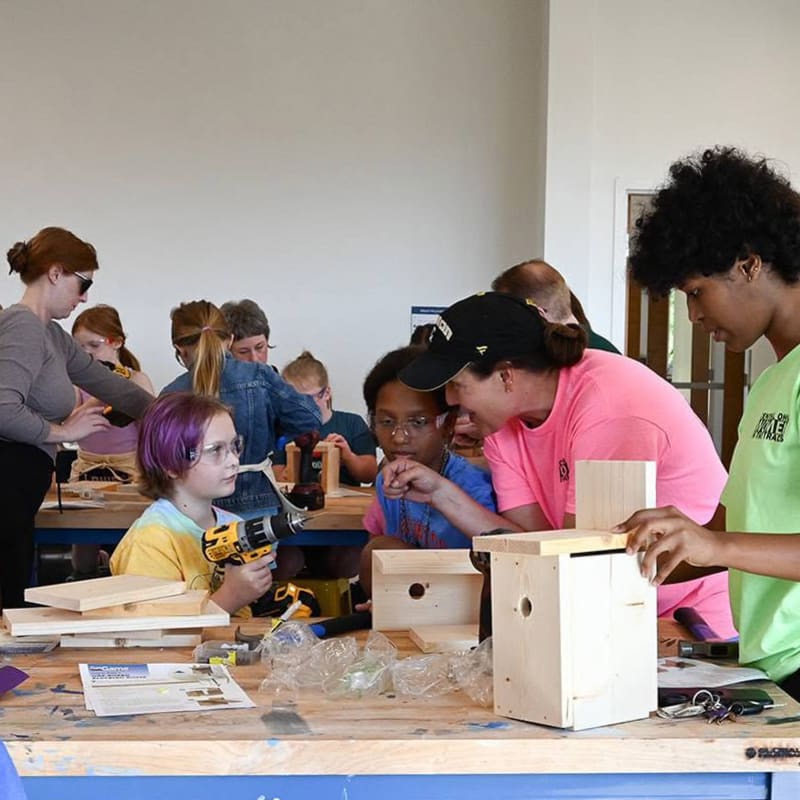 In the foreground, children and adults are gathered around a workbench, building birdhouses with tools, while in the background, more people are engaged in the same activity in a brightly lit room.