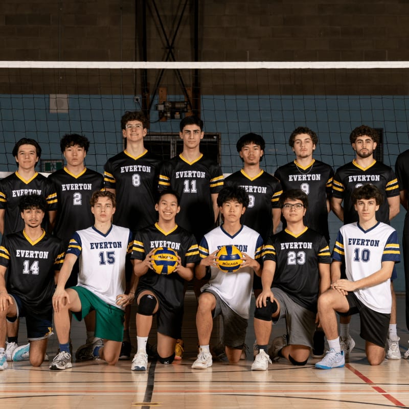 A group of young men in volleyball uniforms pose for a photo in a gymnasium, with a volleyball net visible in the background.