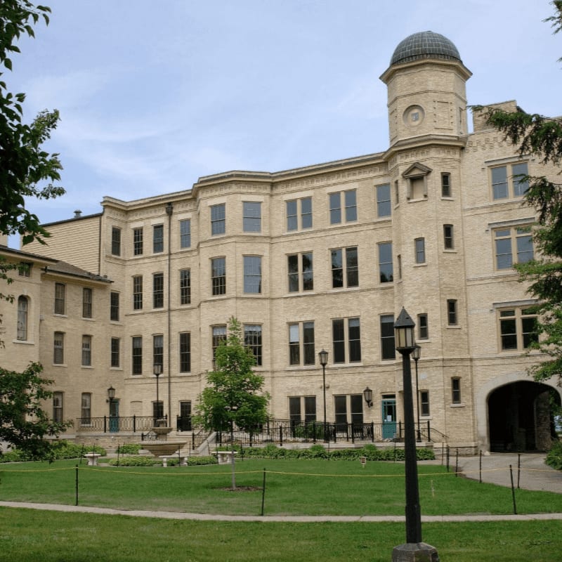 A large, light-colored building with a domed tower stands behind a grassy courtyard with trees and a lamppost in the foreground.