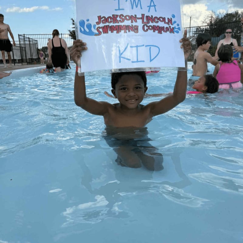 A smiling boy holds a sign in a swimming pool while other people swim in the background.