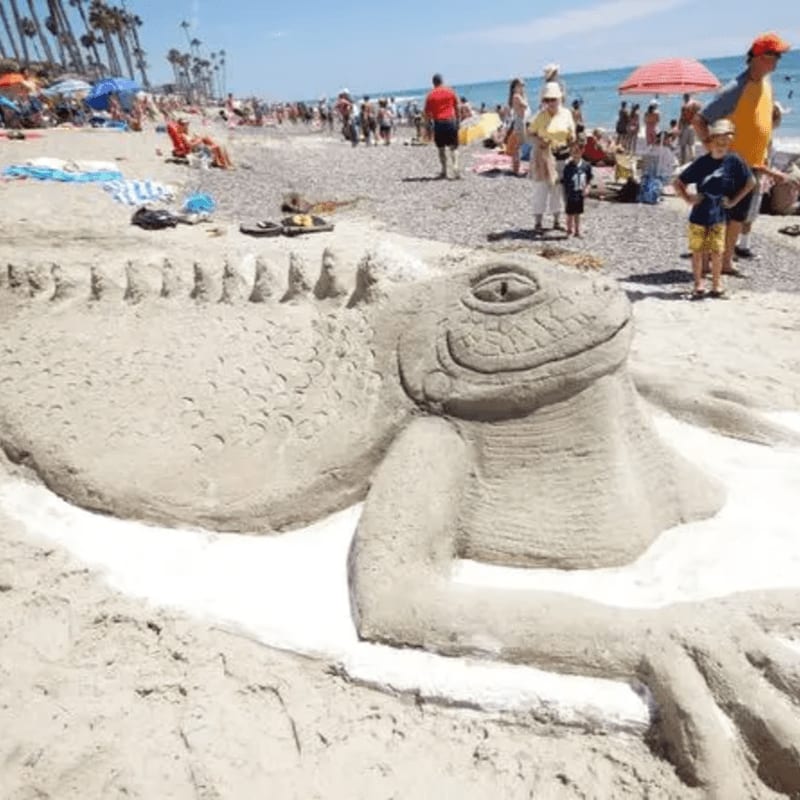 A large sand sculpture of a lizard rests on the beach in the foreground, while a crowded beach with people and umbrellas stretches into the background under a clear blue sky.