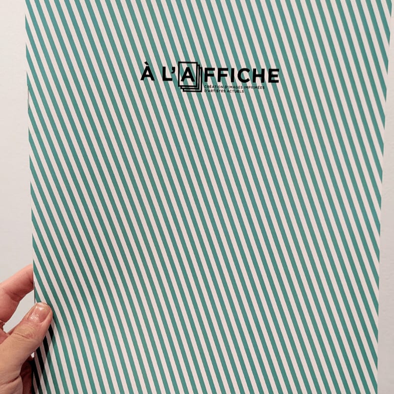 A hand holds a striped green and white paper with the text "À L'AFFICHE" in the upper center against a plain background.