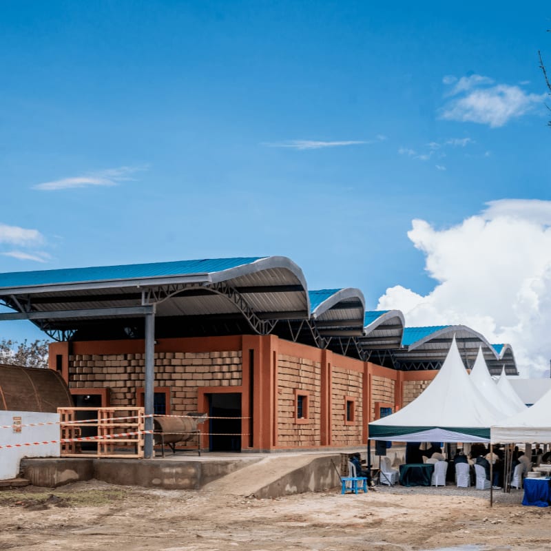 A modern building with a curved metal roof stands in the foreground, while tents and a clear blue sky with scattered clouds form the background.