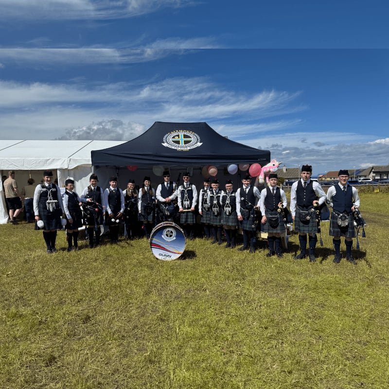 A pipe band in kilts stands in a grassy field in front of a tent, with a blue sky and clouds in the background.